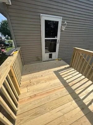 Newly constructed wooden deck with railings attached to home with gray siding and white door